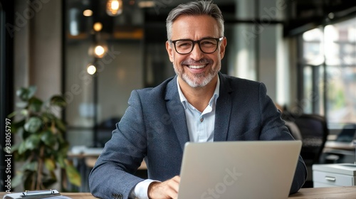 Portrait of happy mid aged business man executive working on laptop in office. Senior company manager investor using computer sitting at desk. Smiling older professional businessman looking at camera