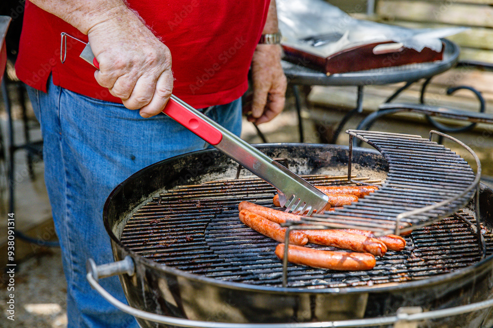 Ταπετσαρία Hot dogs on charcoal kettle grill being turned by older man with tongs