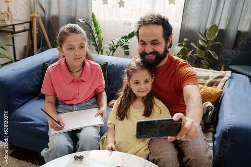 Smiling father sitting on blue couch with daughters capturing selfie. Elder daughter holding notebook and pencil while younger daughter smiling at camera