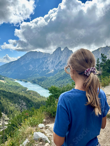 A girl stands at the Estanque viewpoint in the Catalan Pyrenees, taking in the stunning view of a green valley and towering mountains. The dramatic sky adds to the scene's beauty.