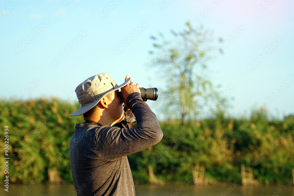 Fototapeta premium Asian man uses binoculars to observe local birds along a river. Soft and selective focus