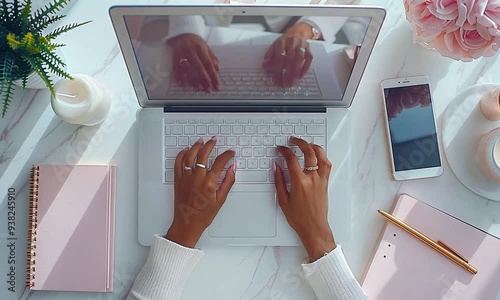 Overhead view of a woman's hands typing on a laptop with a smartphone, notebook, and candles on a marble desk.