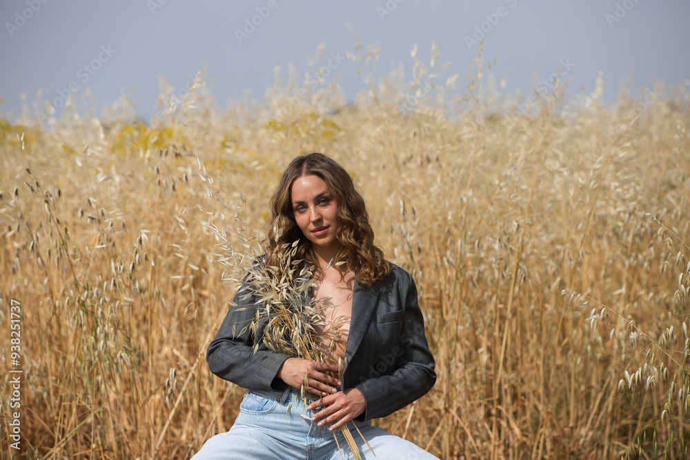 beautiful young blonde woman is dressed in grey jacket, jeans, boots sitting on a stool holding in her hands a bunch of oats from the field where she is the farmer. Concept of sustainable cultivation. beautiful young blonde woman is dressed in grey jacket, jeans, boots sitting on a stool holding in her hands a bunch of oats from the field where she is the farmer. Concept of sustainable cultivation.