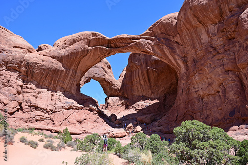 Woman and distant tourists under Double Arch in Arches National Park, Utah on clear sunny summer morning.