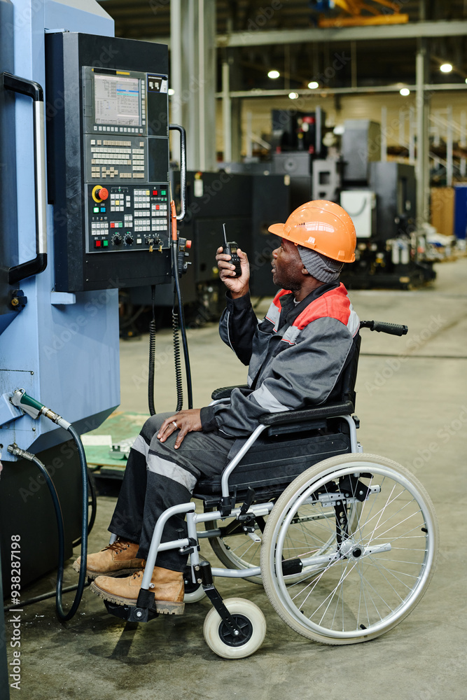 Disabled factory worker operating advanced industrial equipment from wheelchair inside large industrial workspace wearing protective helmet and uniform Disabled factory worker operating advanced industrial equipment from wheelchair inside large industrial workspace wearing protective helmet and uniform