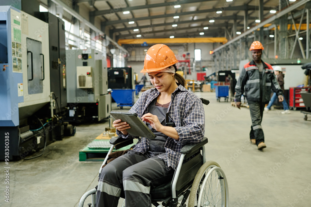 Obraz premium Worker in wheelchair using tablet for managing processes in modern industrial facility. Colleague in background walking between machines, both wearing safety gear
