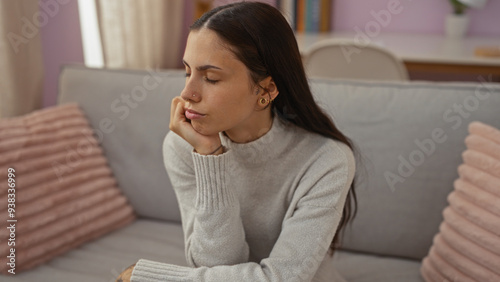 Hispanic woman with brunette hair at home in a living room, wearing a gray sweater and looking thoughtful, with her head rested on her hand against a cozy backdrop. photo