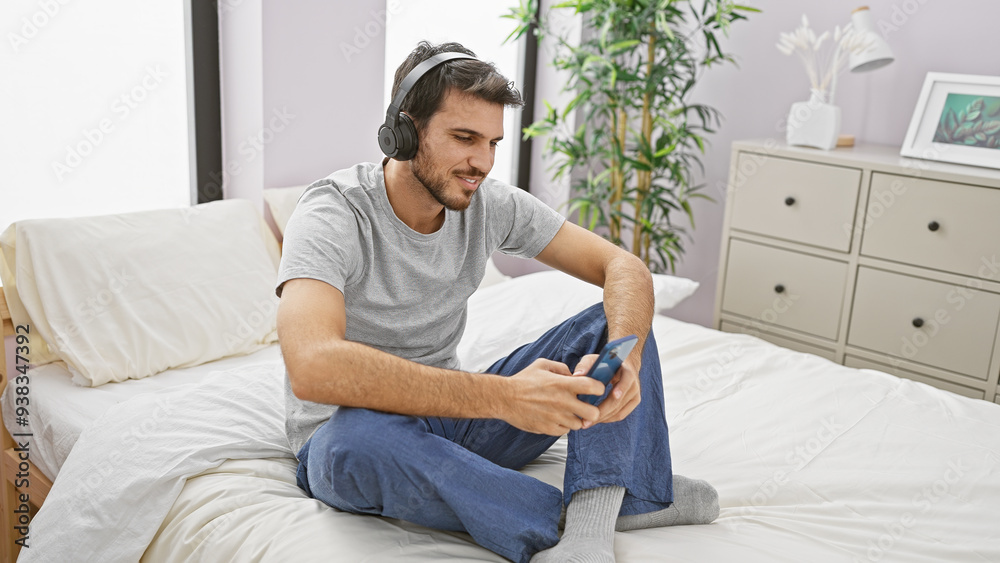 A relaxed hispanic man with a beard enjoys music using headphones and a smartphone while sitting on a bed in a bright bedroom. A relaxed hispanic man with a beard enjoys music using headphones and a smartphone while sitting on a bed in a bright bedroom.