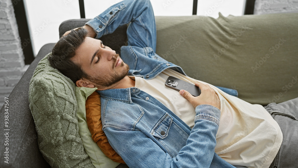 Fototapeta premium A young hispanic man reclining on a sofa with a smartphone in a cozy living room setting, embodying relaxed home life.