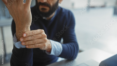 Bearded middle-aged man touching his wrist indoors with blurred office background photo