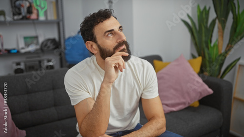 Pensive bearded man in a modern living room thinking and looking upwards, suggesting reflection or decision-making.