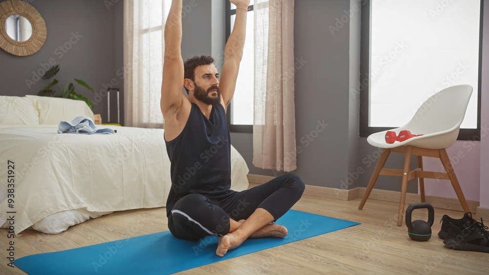 Fototapeta premium A bearded man in a black tank top stretches on a blue yoga mat in a bedroom, near a bed and fitness equipment.