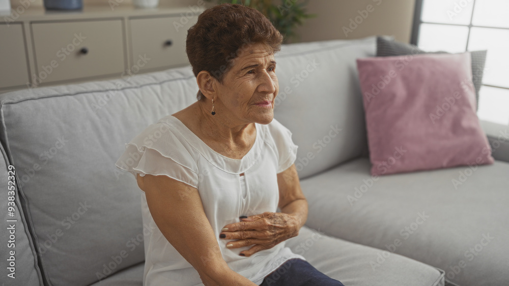Elderly hispanic woman with short hair sitting in a living room, looking pensive and touching her stomach, in a cozy home setting. Elderly hispanic woman with short hair sitting in a living room, looking pensive and touching her stomach, in a cozy home setting.