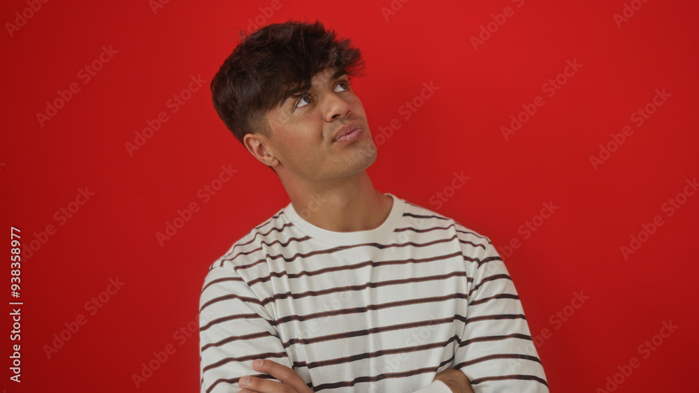 A handsome young hispanic man in a striped shirt crosses his arms and looks upwards against a vivid red background, exuding an introspective and thoughtful expression. A handsome young hispanic man in a striped shirt crosses his arms and looks upwards against a vivid red background, exuding an introspective and thoughtful expression.