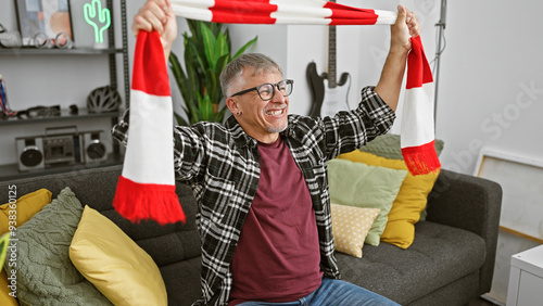 Middle-aged man cheering with a scarf in a cozy living room, showcasing joyful enthusiasm. photo