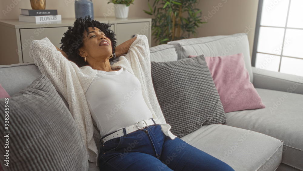 A young african american woman relaxes in a modern living room with cozy decor, wearing casual clothes and sitting comfortably on a sofa with pillows. A young african american woman relaxes in a modern living room with cozy decor, wearing casual clothes and sitting comfortably on a sofa with pillows.