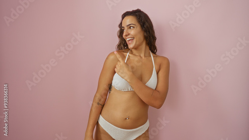 Beautiful young hispanic woman in a white bikini smiling and pointing to the side over an isolated pink background