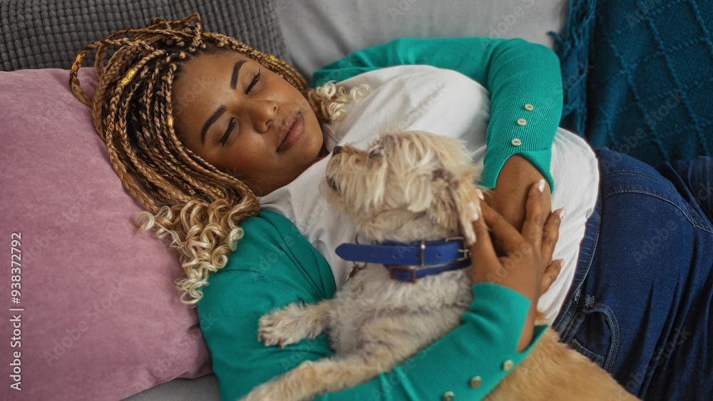 A young african american woman with braids is relaxing indoors on a couch in her living room, cuddling her small dog closely. A young african american woman with braids is relaxing indoors on a couch in her living room, cuddling her small dog closely.