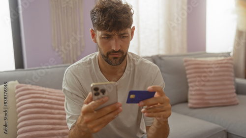 Fotografie Young man sitting in a living room holding a phone and credit card, looking concerned, possibly about online shopping or banking issues, with cozy home furnishings visible in the background