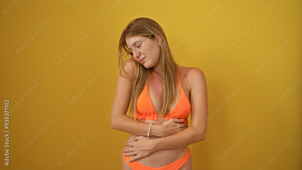Young woman with blonde hair in an orange bikini holding her stomach in pain, isolated against yellow background Young woman with blonde hair in an orange bikini holding her stomach in pain, isolated against yellow background