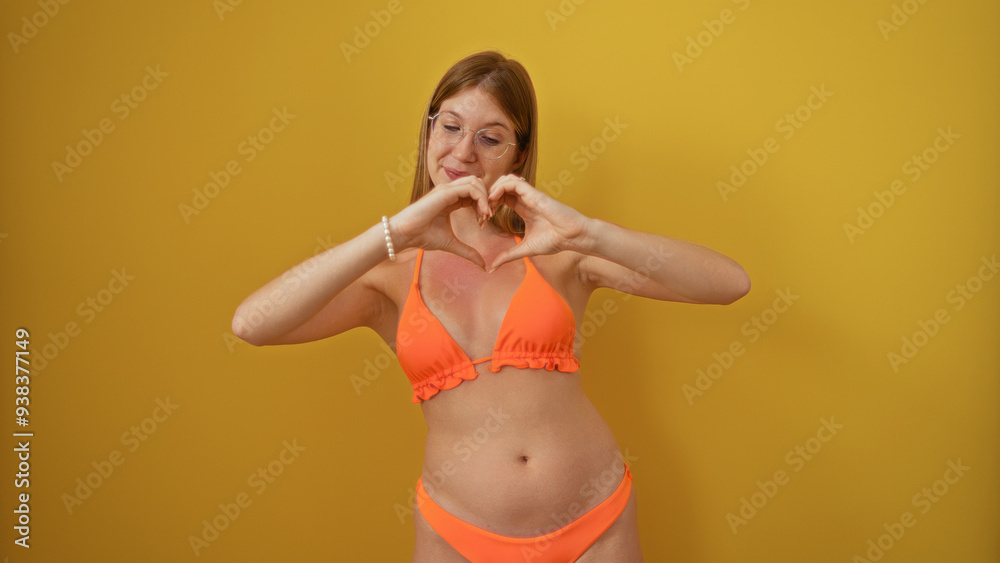 Naklejka premium Young woman in a bikini making a heart shape with hands over an isolated yellow background, highlighting her blonde hair and attractive smile.
