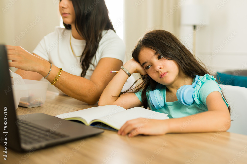 Unmotivated elementary school girl feeling bored while studying with her mother and looking sad at the kitchen table Unmotivated elementary school girl feeling bored while studying with her mother and looking sad at the kitchen table