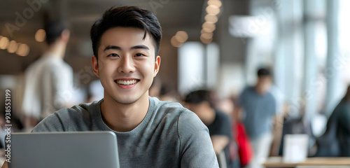 A handsome young Asian man with cool hair style is smiling while sitting at a desk in front of his laptop, dressed casually