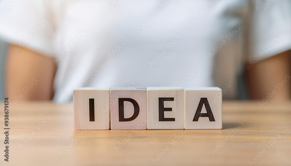 Naklejka premium 木製のブロックに「IDEA」という文字が書かれており、それがテーブルの上に並べられています。背景には白いシャツを着た人物がぼんやりと写っており、アイデアの概念や創造性を表現しています。全体的にシンプルでフォーカスが「IDEA」に当てられています。generative AI