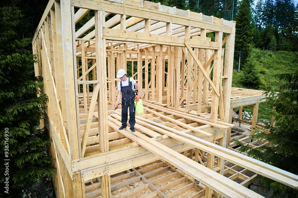 Laborer constructing wooden frame house near forest. Man treating woods, applying fire retardant using sprayer, while dressed in protective suit, helmet. Concept of modern eco-friendly construction. Laborer constructing wooden frame house near forest. Man treating woods, applying fire retardant using sprayer, while dressed in protective suit, helmet. Concept of modern eco-friendly construction.