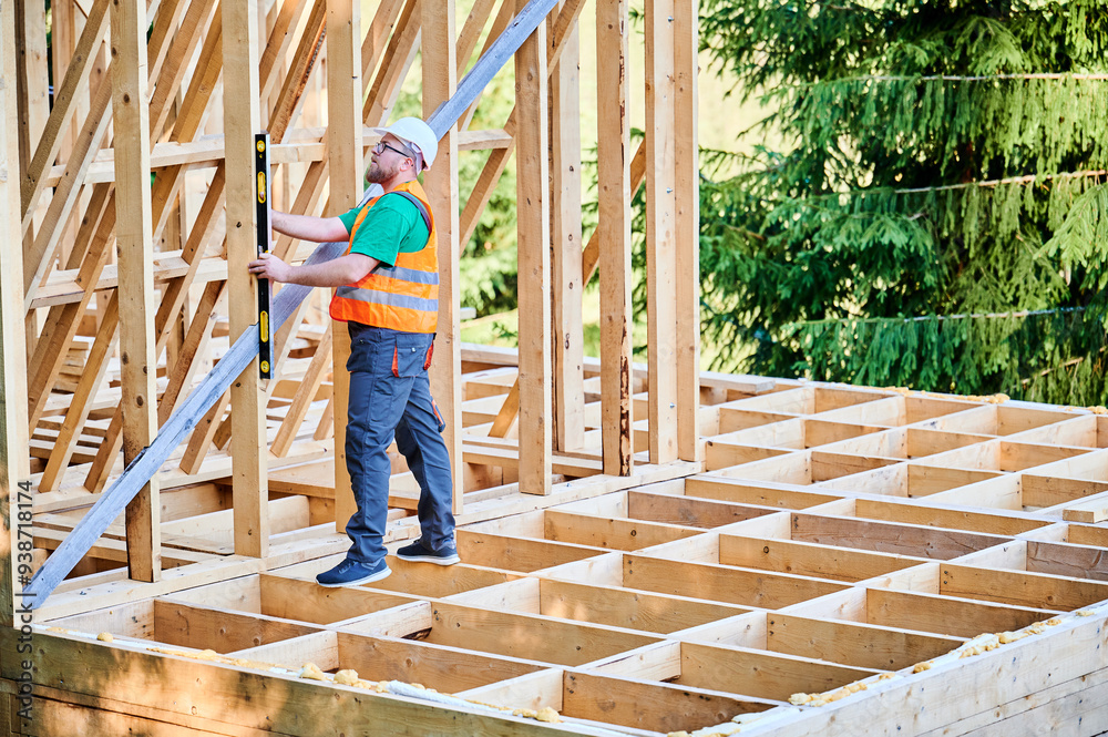 Carpenter constructing two-storey wooden frame house. Man inspects walls for levelness using spirit level, wearing protective overalls, helmet and orange vest. Concept of eco-friendly construction. Carpenter constructing two-storey wooden frame house. Man inspects walls for levelness using spirit level, wearing protective overalls, helmet and orange vest. Concept of eco-friendly construction.