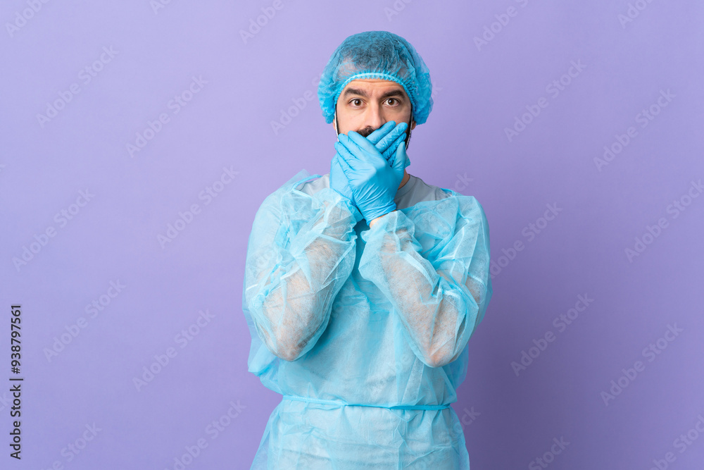 Surgeon man with beard with blue uniform over isolated purple background covering mouth with hands Surgeon man with beard with blue uniform over isolated purple background covering mouth with hands