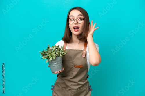 Gardener Ukrainian woman holding a plant isolated on blue background listening to something by putting hand on the ear