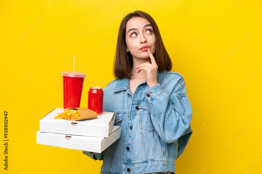 Young Ukrainian woman holding fast food isolated on yellow background having doubts while looking up Young Ukrainian woman holding fast food isolated on yellow background having doubts while looking up