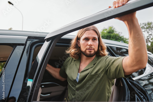 Young man with long hair getting out of a car in a casual outfit, surrounded by greenery