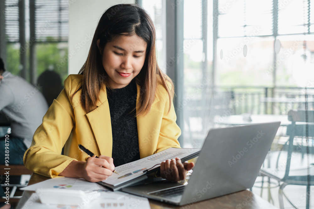 Focused young woman in a yellow blazer working on a laptop and taking notes in a bright, modern office setting.