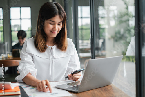 A young woman in a white blouse works on her laptop and smartphone at a modern office desk, focusing on her tasks.