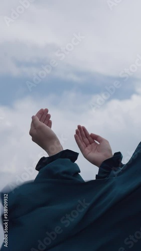 Vertical Screen: Muslim Woman in a Fluttering Dress Praying with Hands Raised Outdoors Under the Cloudy Sky. Concept of Islam, faith, spirituality, hope, worship, dua, belief, religion