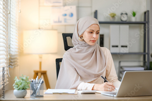 A woman wearing a head scarf sits at a desk with a laptop and a pen