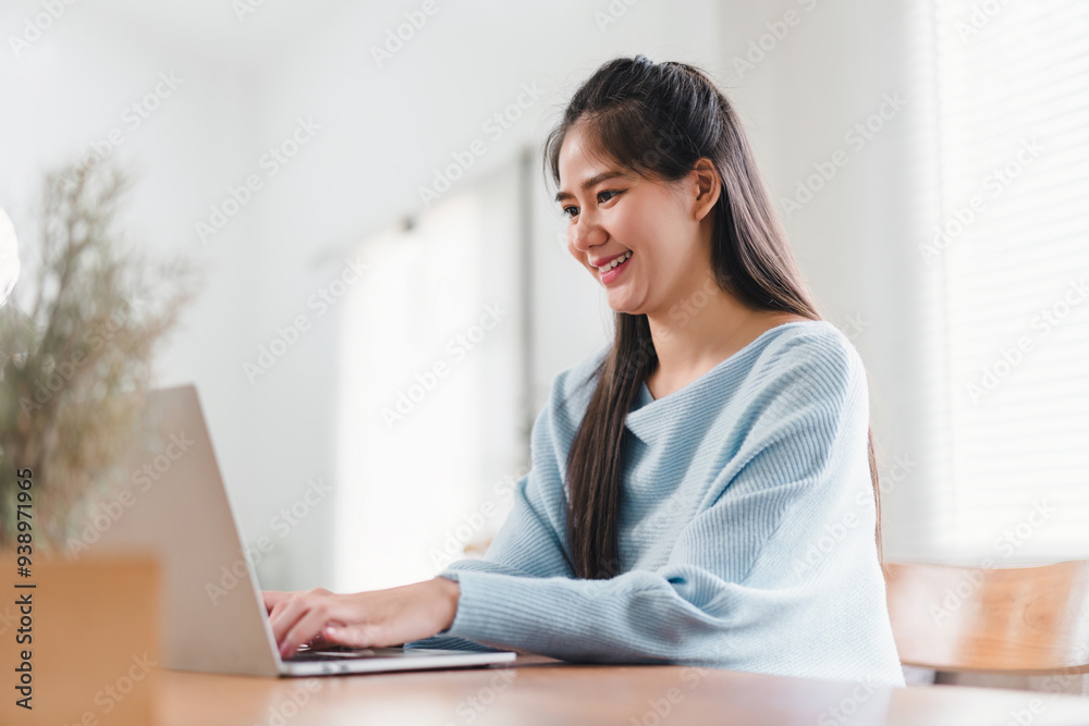 Smiling Asian woman typing on laptop while sitting at home, focused on work or study in a comfortable setting. Smiling Asian woman typing on laptop while sitting at home, focused on work or study in a comfortable setting.