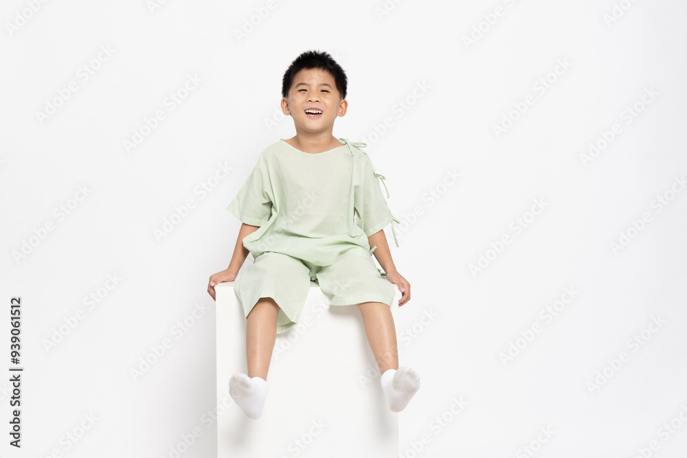 Happy Asian little boy in hospital patient uniform sitting on white box isolated on white background Happy Asian little boy in hospital patient uniform sitting on white box isolated on white background