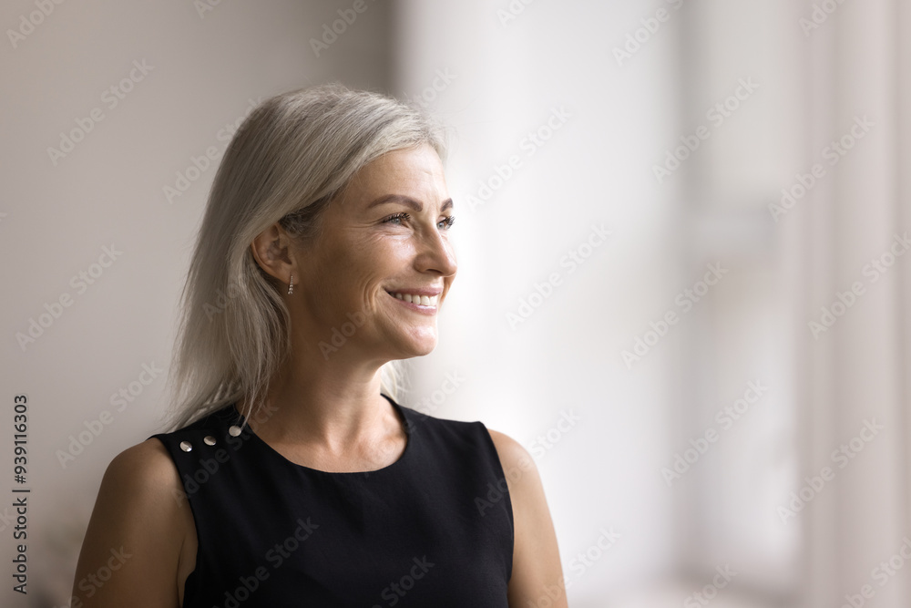 Head shot beautiful middle-aged female smiling looking out window, standing alone in modern living room, admire view from apartment, deep in pleasant thoughts, recollect, enjoy warm memories at home Head shot beautiful middle-aged female smiling looking out window, standing alone in modern living room, admire view from apartment, deep in pleasant thoughts, recollect, enjoy warm memories at home