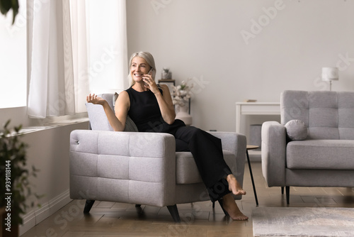 Cheerful middle-aged woman relax on comfortable armchair, talk on smartphone to friend, have pleasant conversation, share information, enjoy distancing communication. Wireless technology, connection