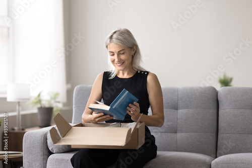 Smiling mature woman sitting on sofa unpacking box, get package, opening received parcel with ordered books using easy and comfortable on-line bookshop delivery services. Hobby, leisure, book lover