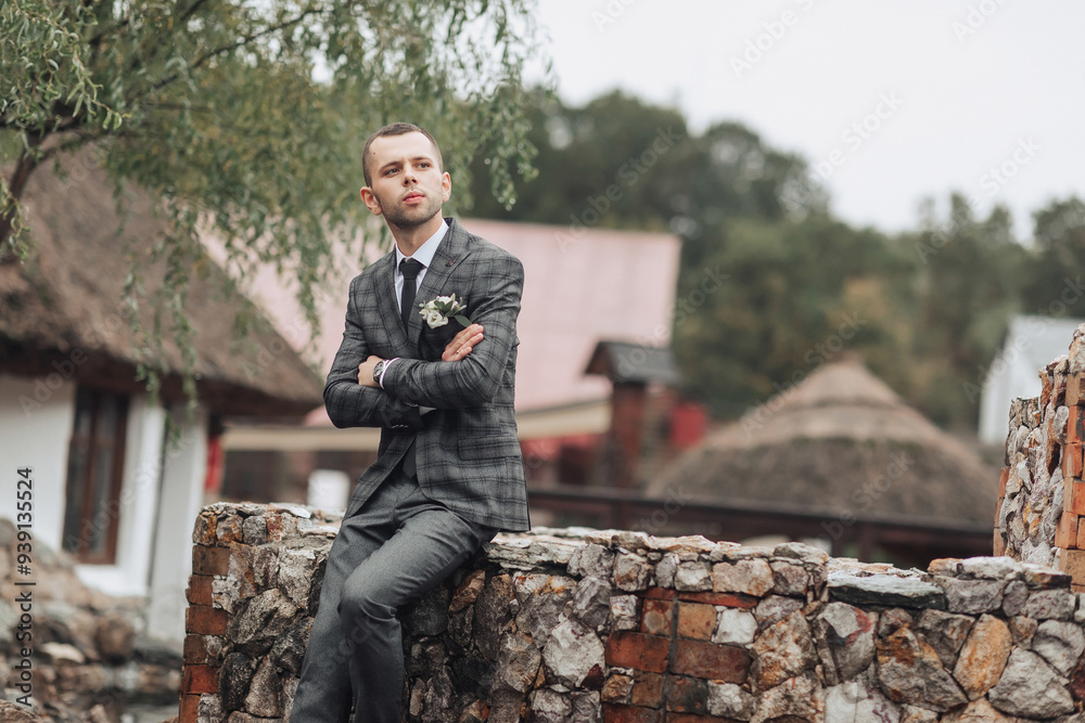 Fototapeta premium A man in a suit is sitting on a stone wall. He is wearing a tie and a flower in his lapel. The man is looking off into the distance