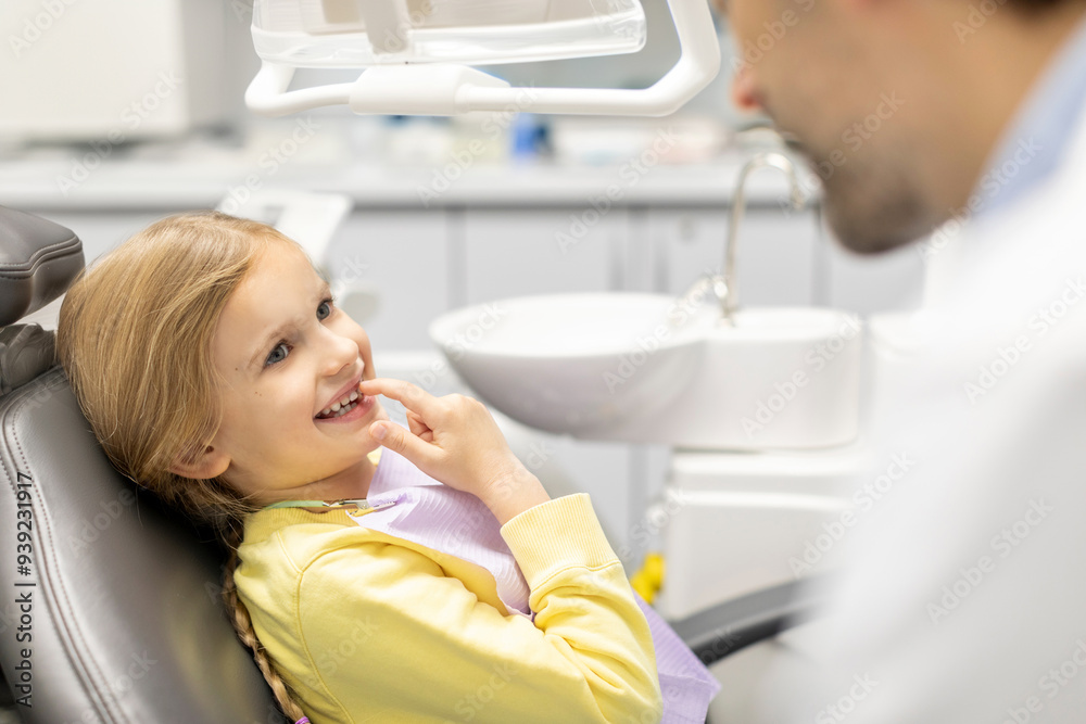 Fototapeta premium Adorable little girl showing her baby teeth to male pediatrics dentist and pointing to her smile, sitting in chair in clinic office