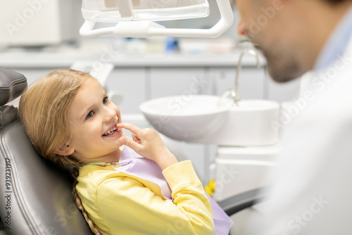 Adorable little girl showing her baby teeth to male pediatrics dentist and pointing to her smile, sitting in chair in clinic office