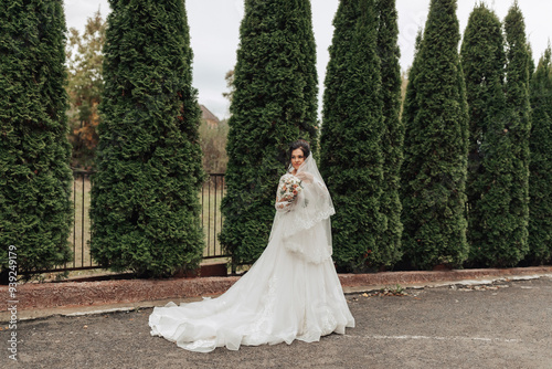 A woman in a white dress stands in front of a row of trees. She is holding a bouquet of flowers and a veil. The scene is serene and peaceful, with the woman looking into the distance