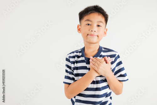 Happy young kid boy in casual clothes holding hands at chest close to heart meditation and smiling in studio short isolated on white background, primary child believe faith and love gratitude