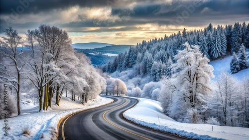 Winter landscape featuring a snowy asphalt road winding through a frozen forest, surrounded by bare trees and rolling hills under a grey sky. photo