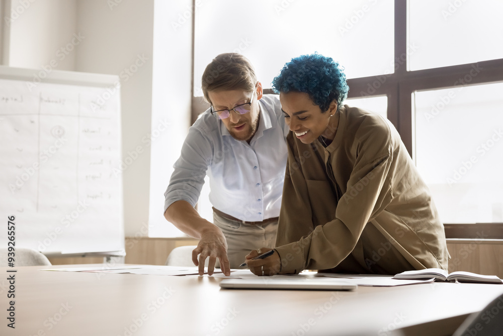 Fototapeta premium Two young multinational colleagues standing at desk, examining printed documents, discussing and analyzing infographic data working on financial statement and report, preparing project or presentation